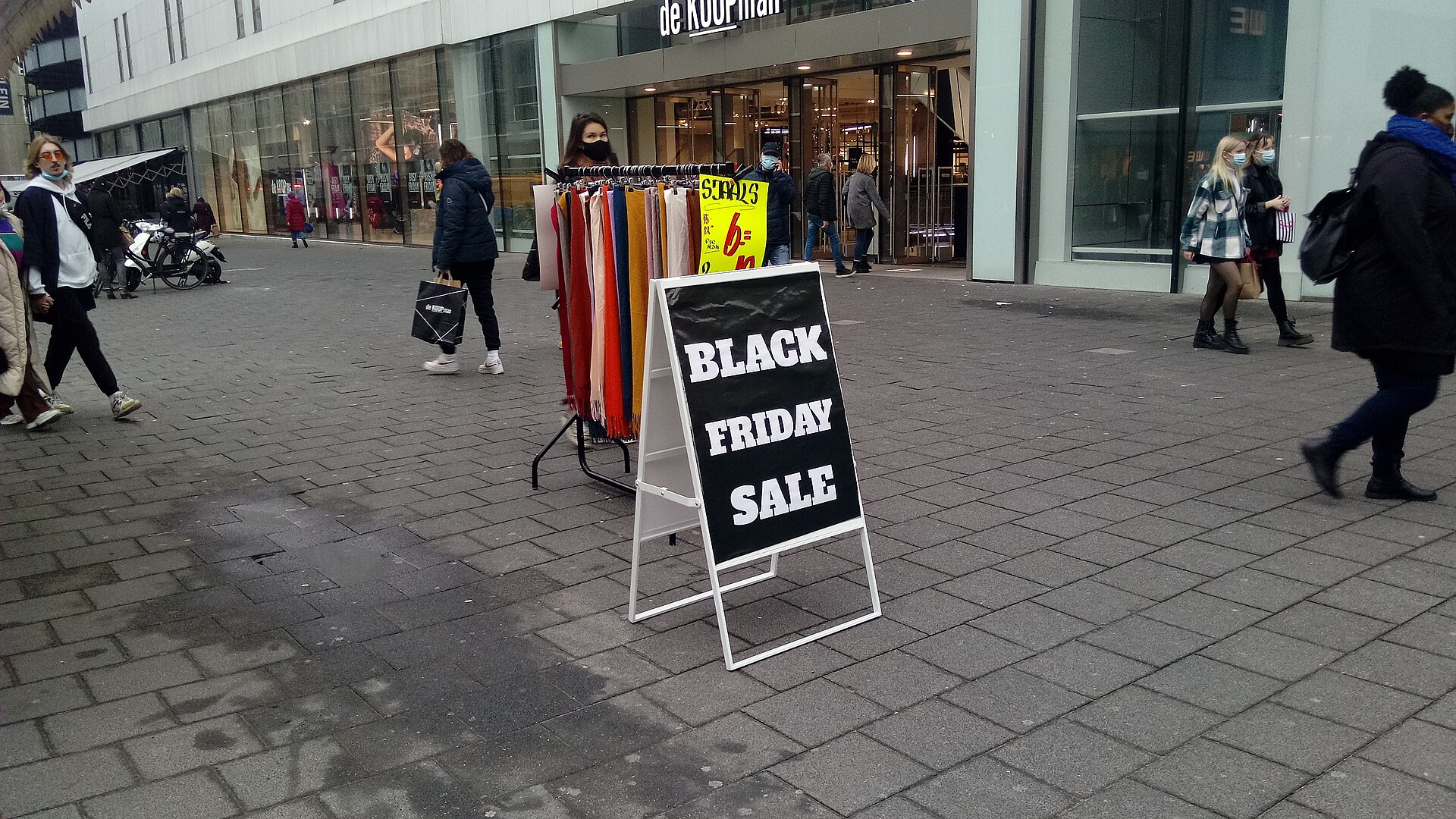 Schaufenster mit großem Rabattschild, dahinter Kleiderstangen und Kunden im hellen Ladeninneren