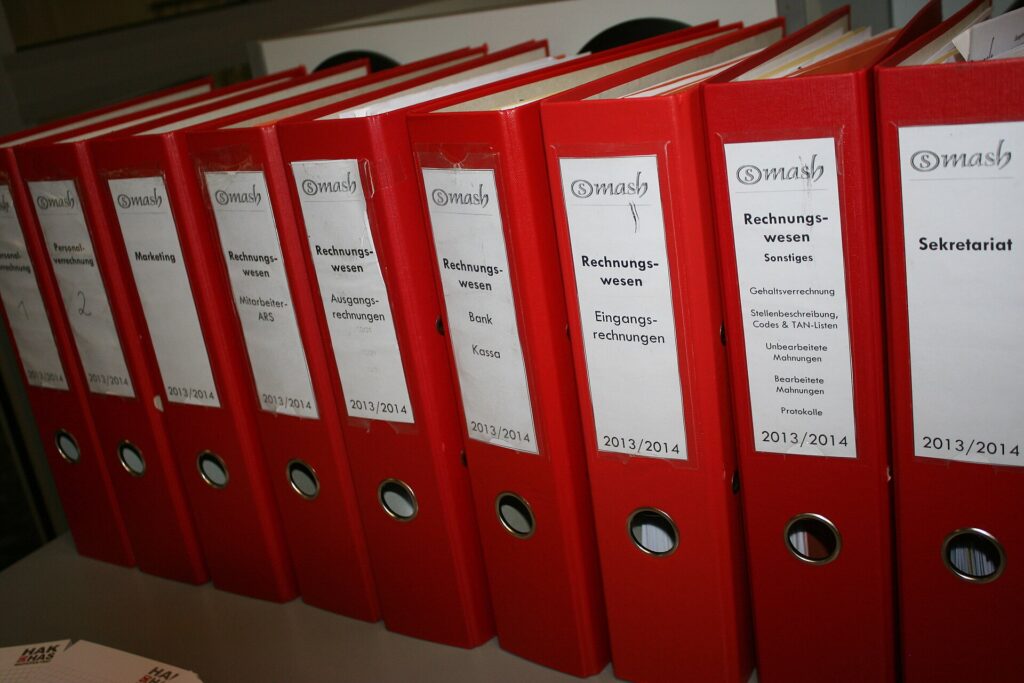 Regal mit beschrifteten Ordnern, davor steht eine geöffnete Ablagebox aus Pappe