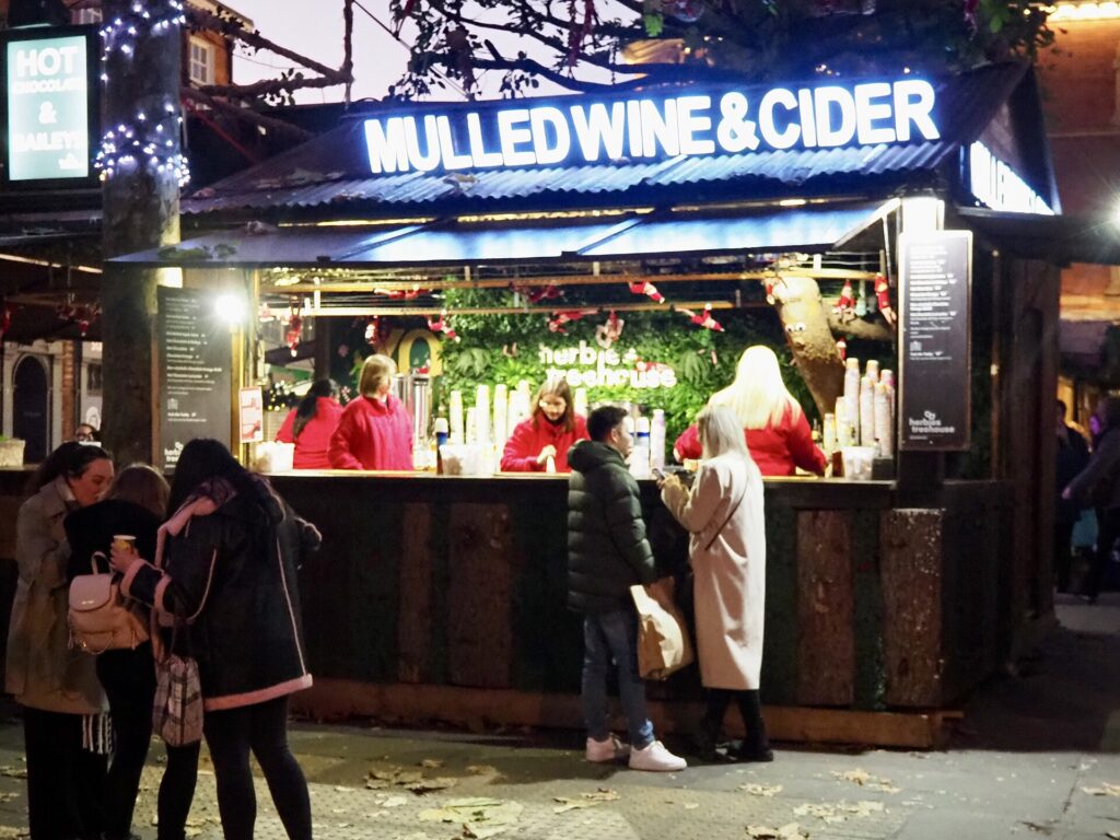 Beleuchteter Stand auf einem Weihnachtsmarkt am Abend, davor stehen mehrere Besucher