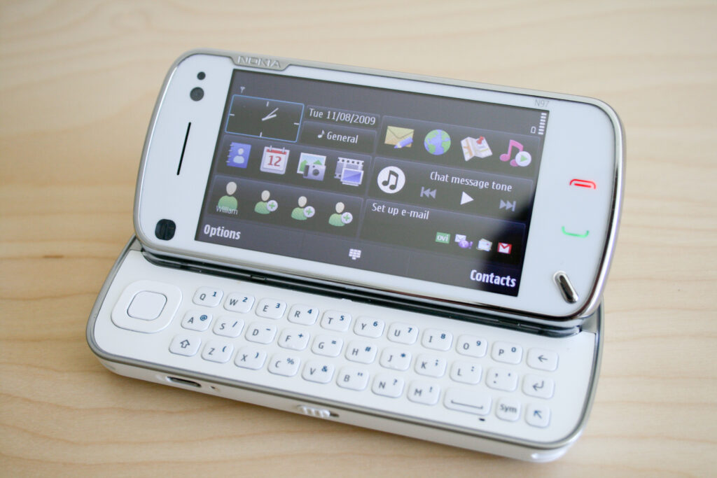 Hand hält ein Smartphone mit eingeblendeter Bildschirmtastatur über einem hellen Schreibtisch
